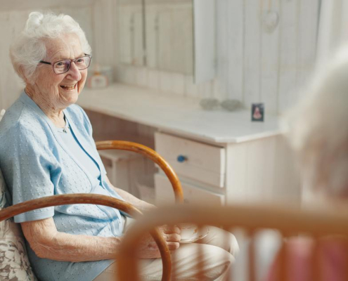 An elderly woman with dementia care at home, smiling warmly while seated in a rocking chair in a comfortable, sunlit room