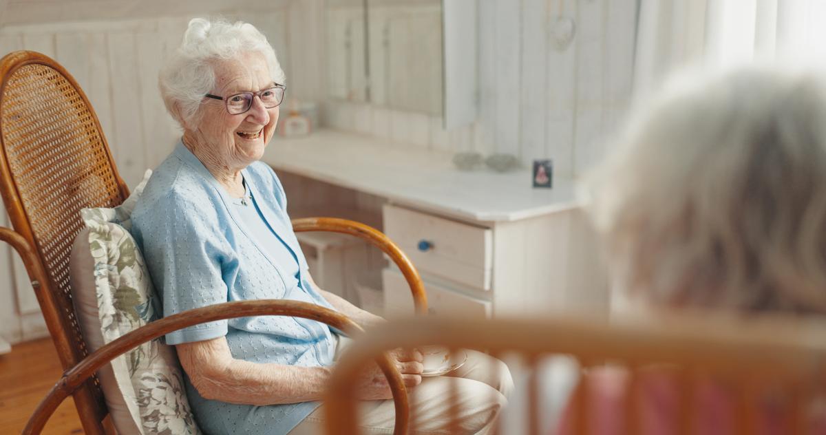 dementia-care-at-home-senior-woman-smiling An elderly woman with dementia care at home, smiling warmly while seated in a rocking chair in a comfortable, sunlit room